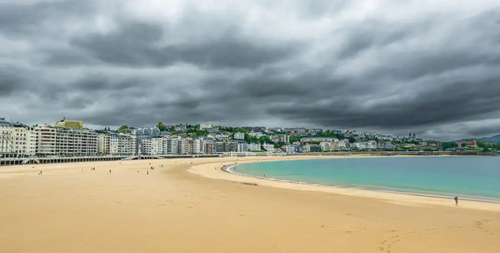 Dramatic cloudy sky over La Concha Beach, San Sebastián, with urban buildings lining the shore.